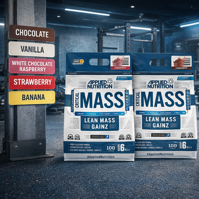 Two packages of Applied Nutrition Critical Mass supplement in a gym setting with flavor options displayed.