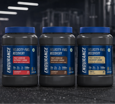 Three bottles of Endurance Velocity-Fuel Recovery Drink mix on a dark surface with a blurred gym background.