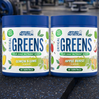 Two containers of Applied Nutrition Critical Greens on a gym floor with exercise equipment in the background.