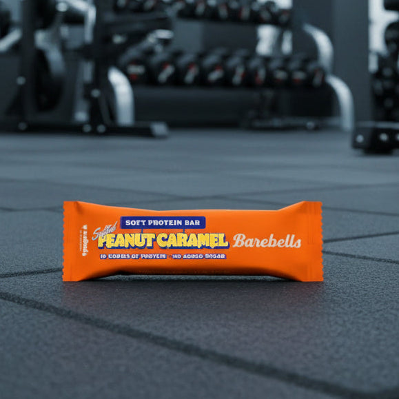 Peanut Caramel Barebells protein bar on a gym floor with blurred gym equipment in the background