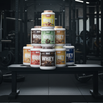 Stack of various flavors of NXT Nutrition Whey protein containers in a gym setting.