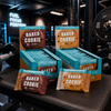 MyProtein Baked Cookie packages displayed on a gym bench with gym equipment in the background.