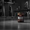 Container of NXT Nutrition Beef Protein Isolate with cola flavour on a gym background