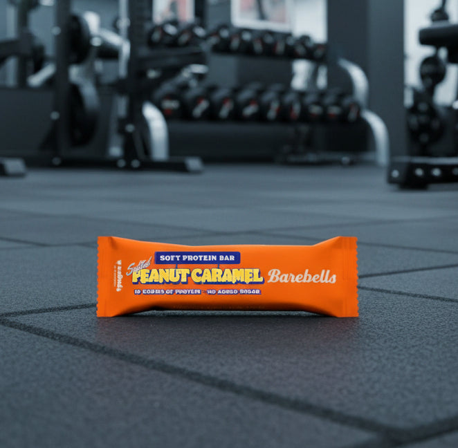 Peanut Caramel Barebells protein bar on a gym floor with blurred gym equipment in the background